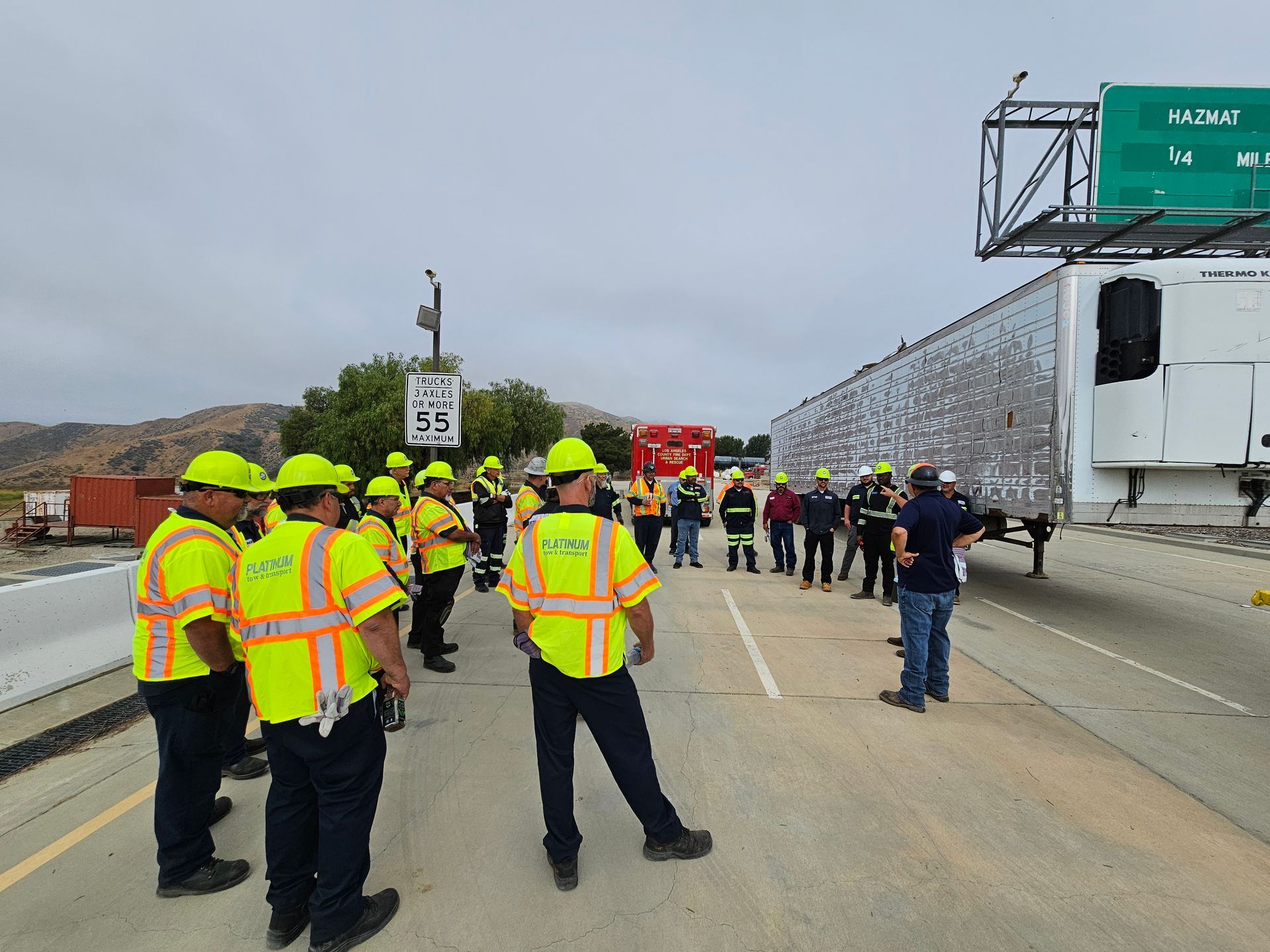 Workers in neon safety vests gather on a highway beside a damaged truck and a stopped trailer.