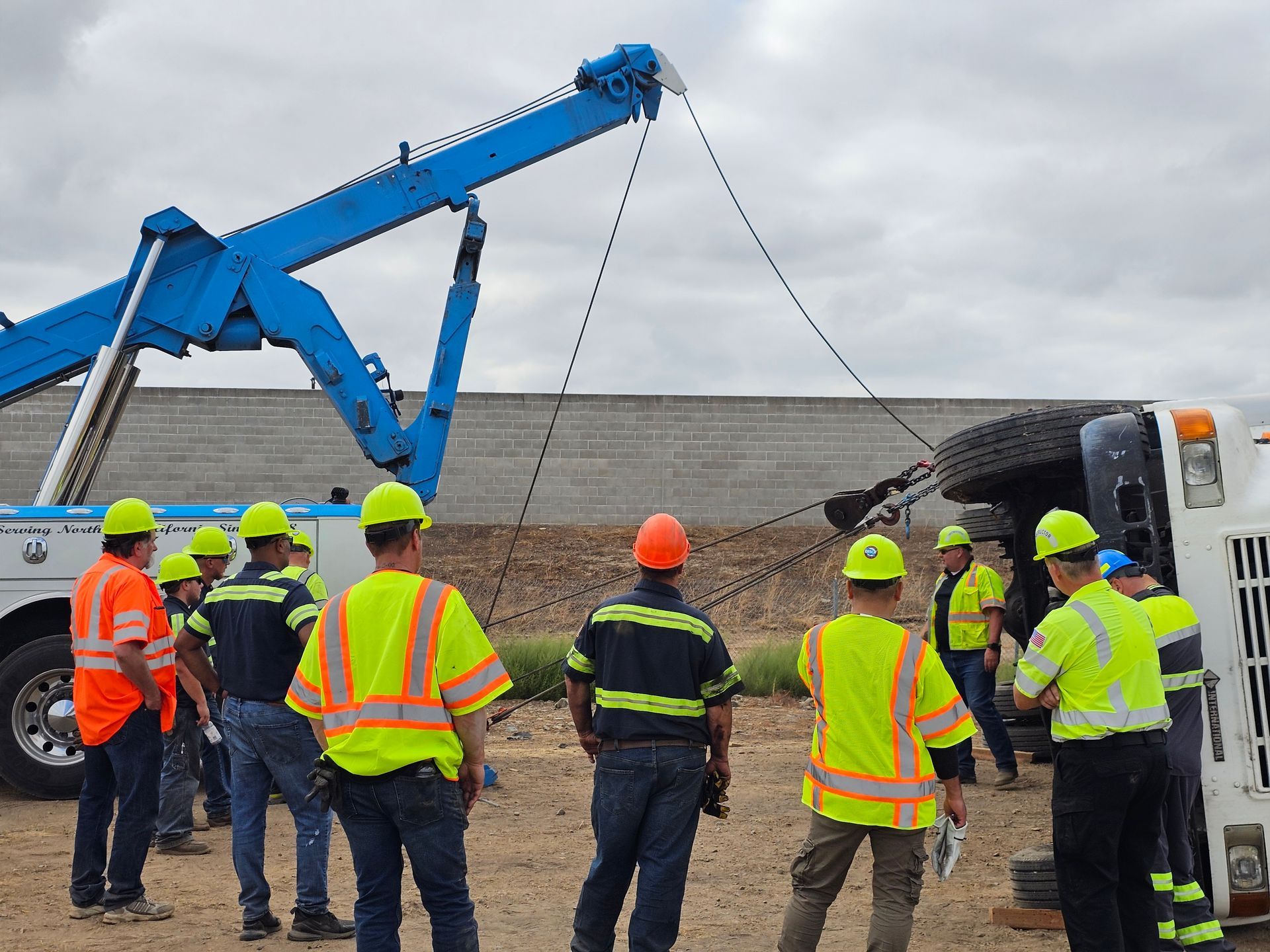 Workers in high-visibility gear gather around a blue crane lifting a truck bed on a roadside.