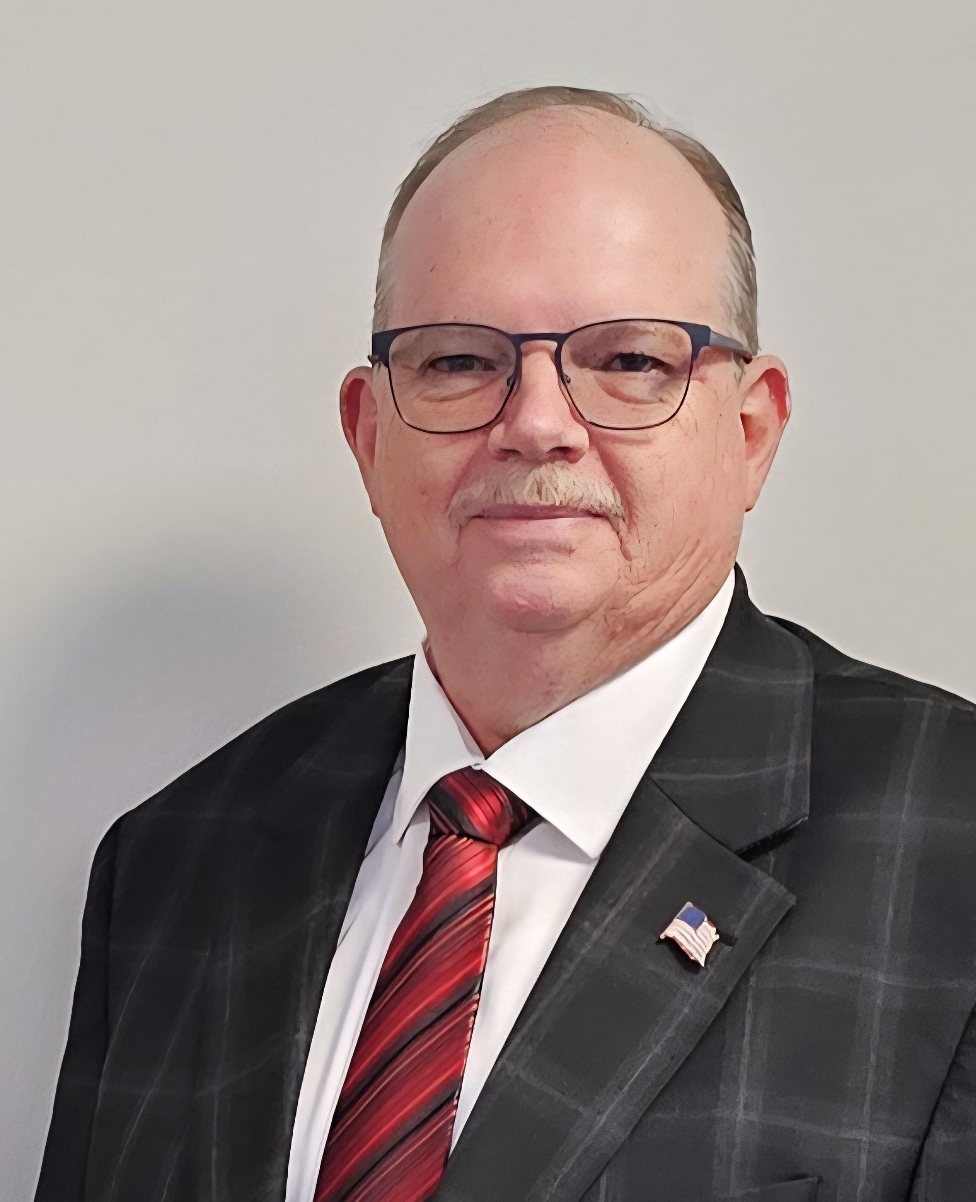 Portrait of a man in a dark suit, red striped tie, and glasses against a plain gray background