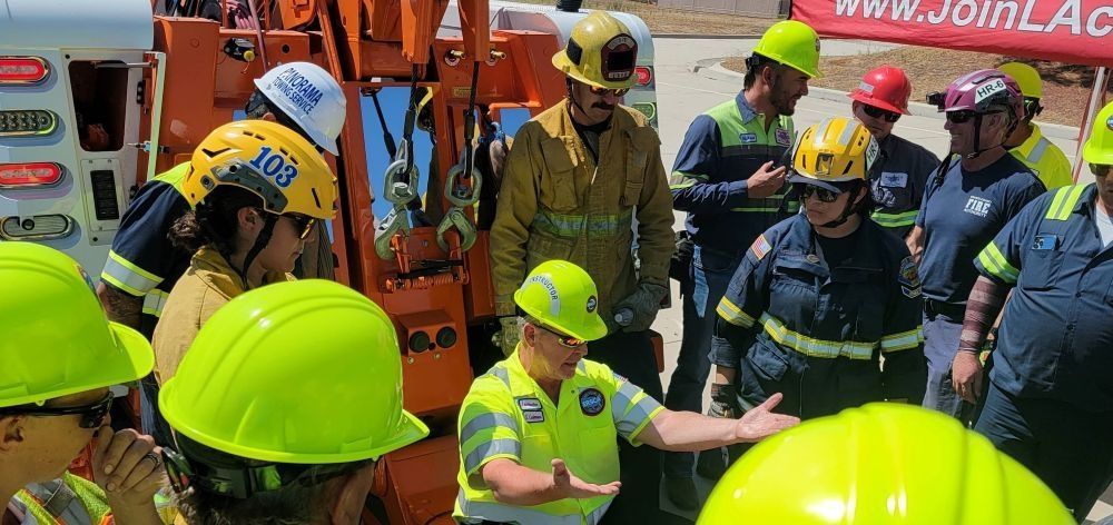 Firefighters and emergency workers in helmets gather at an incident scene near an orange truck.