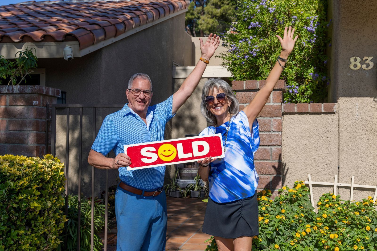 House with FOR SALE sign on front lawn. Cream-colored siding, blue sky.