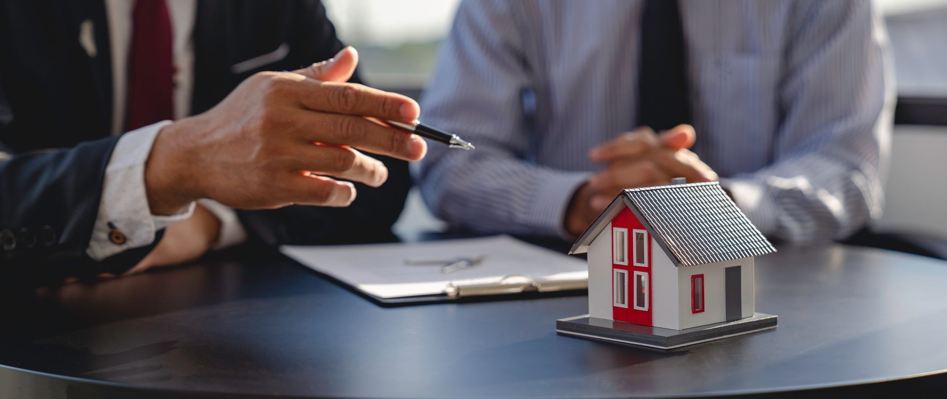Two people at a table reviewing paperwork with a model house. One points with a pen.