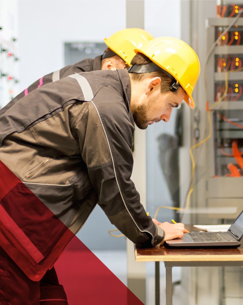 Worker in yellow hard hat leaning over a laptop at a workbench in an industrial setting