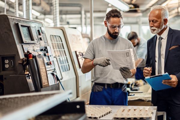 Two workers reviewing paperwork beside industrial equipment in a factory.