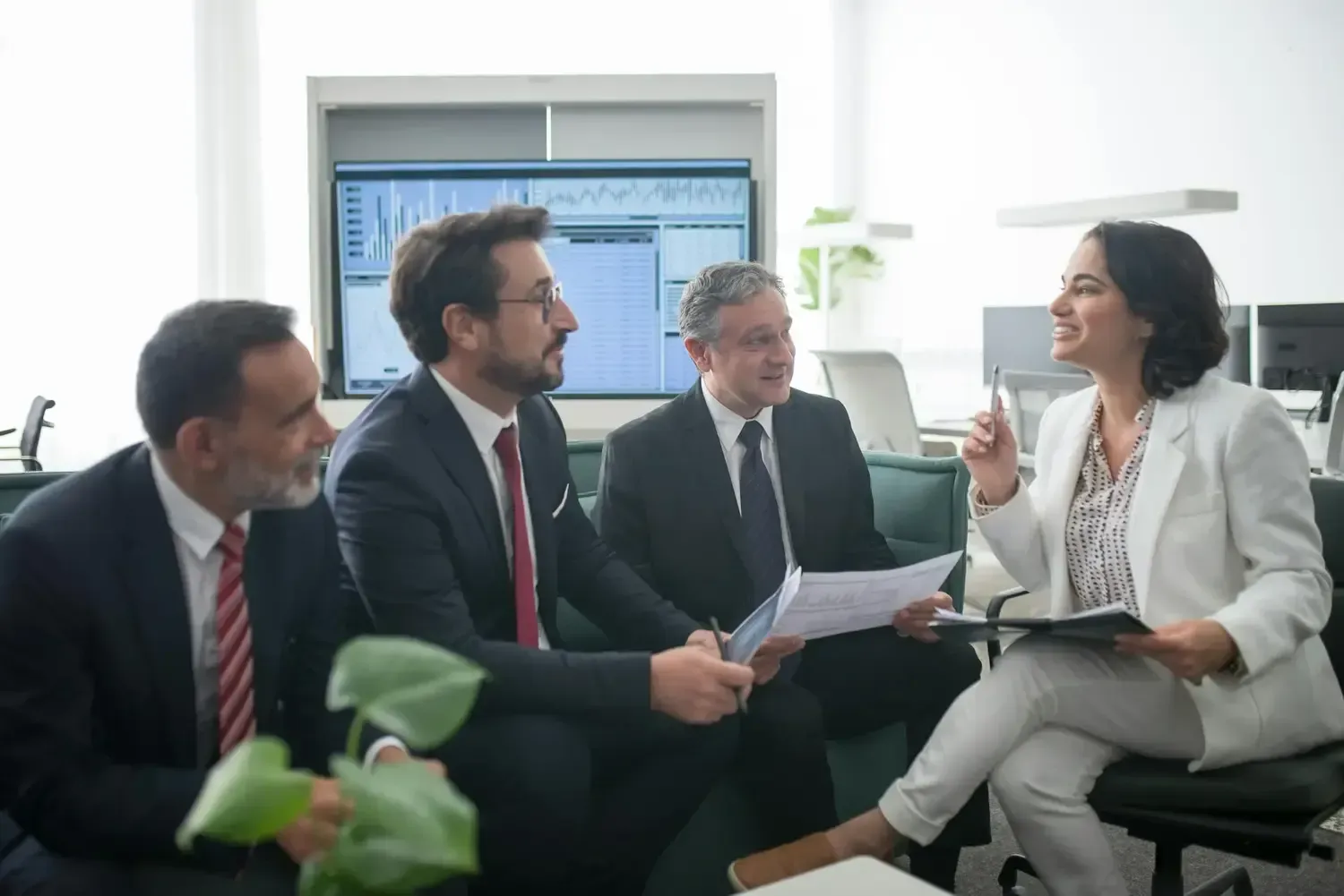 Four businesspeople in suits discussing documents in a bright office meeting room