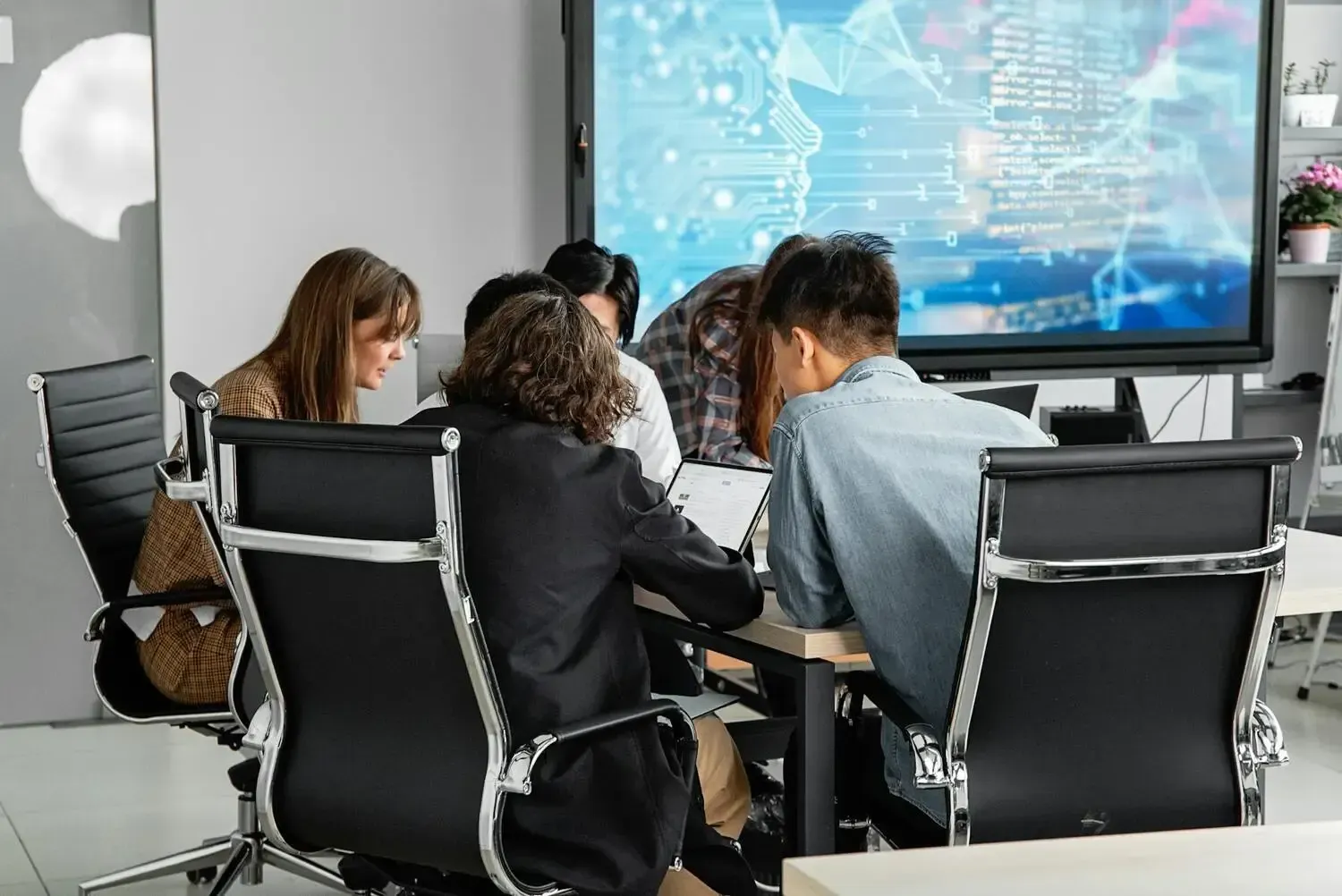 Group seated in a meeting room, facing a presentation screen with charts and data.