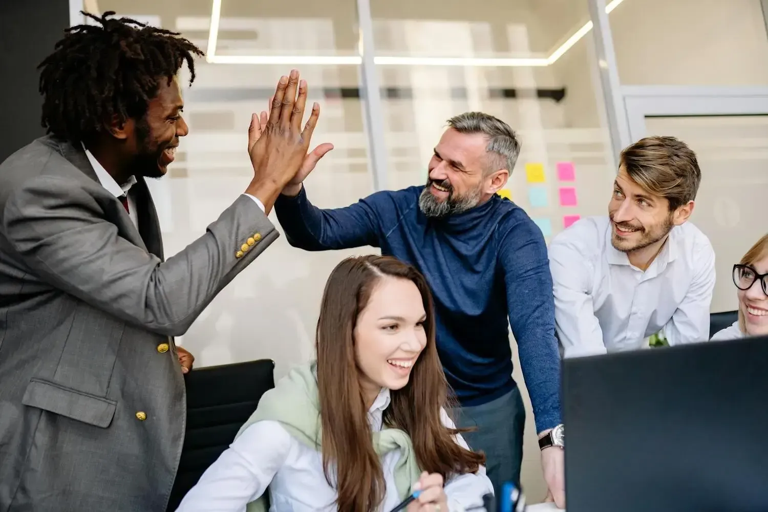 Colleagues in an office high-five while a woman smiles at a desk nearby