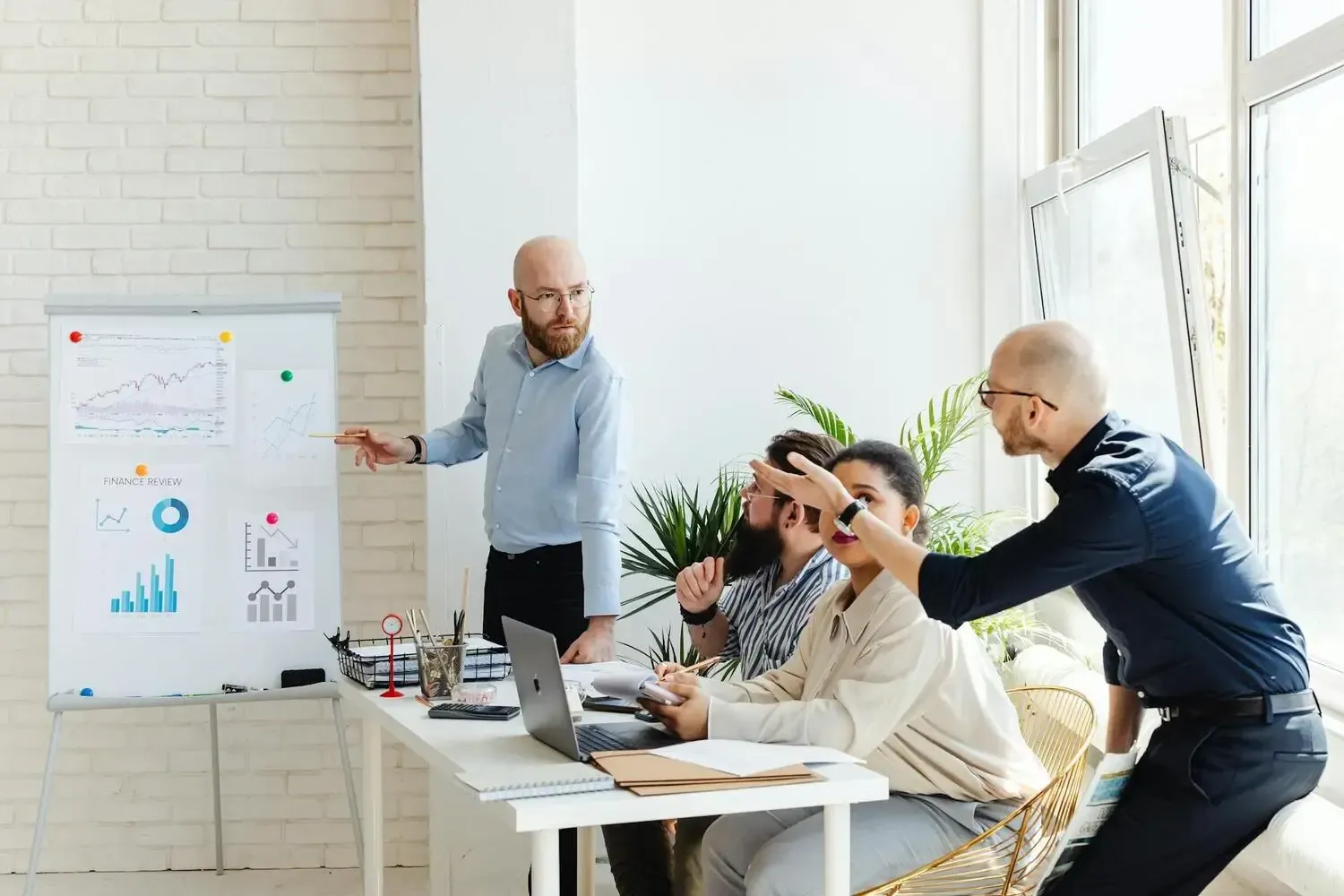 Team meeting in a bright office, with three people discussing charts by a whiteboard and laptop.