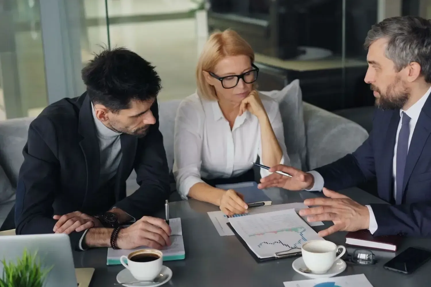 Three coworkers discussing documents at a conference table with coffee cups in a glass-walled office.