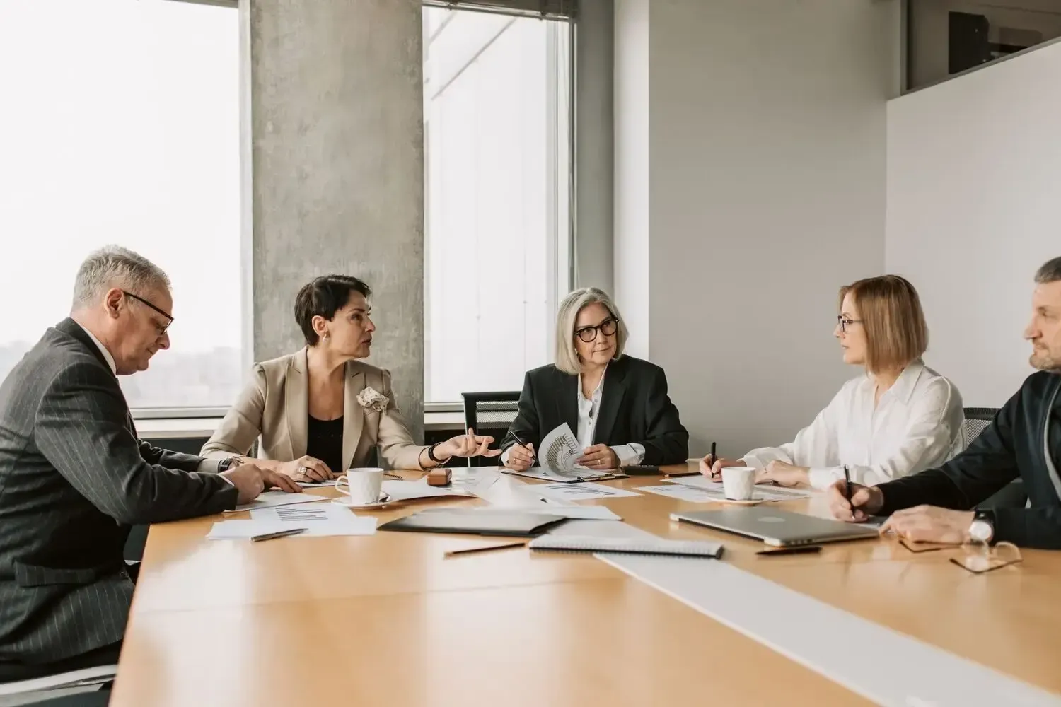 Business meeting around a conference table in a bright office, with documents and laptops spread out.