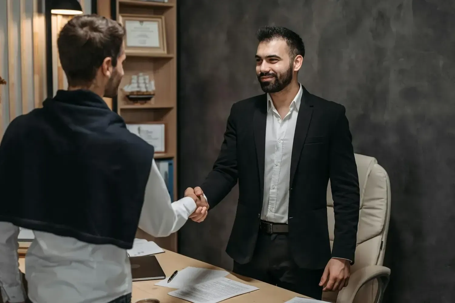 Two people shaking hands in an office, one standing behind a desk and smiling