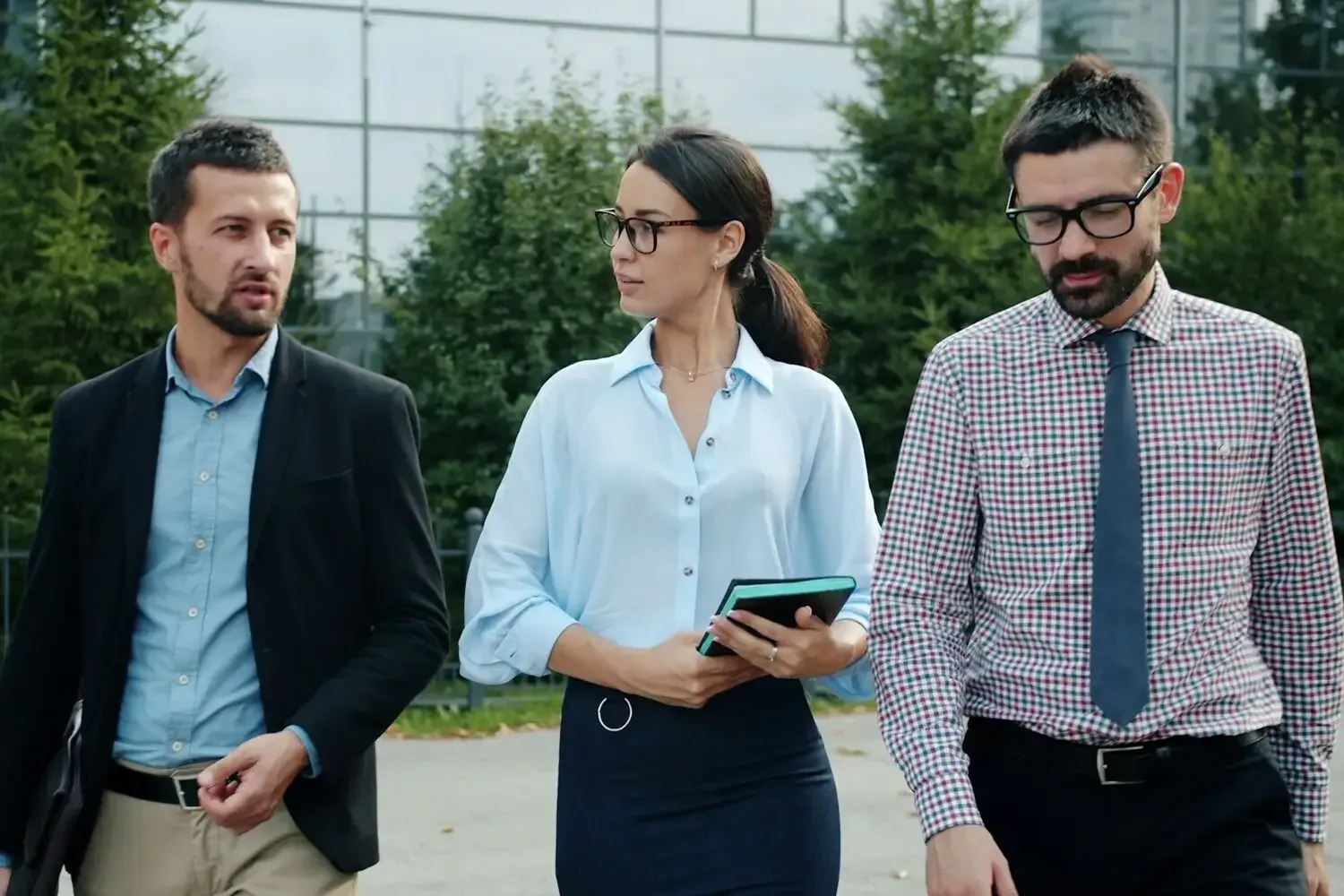 Three professionals walking outdoors by a modern glass building, one holding a tablet.
