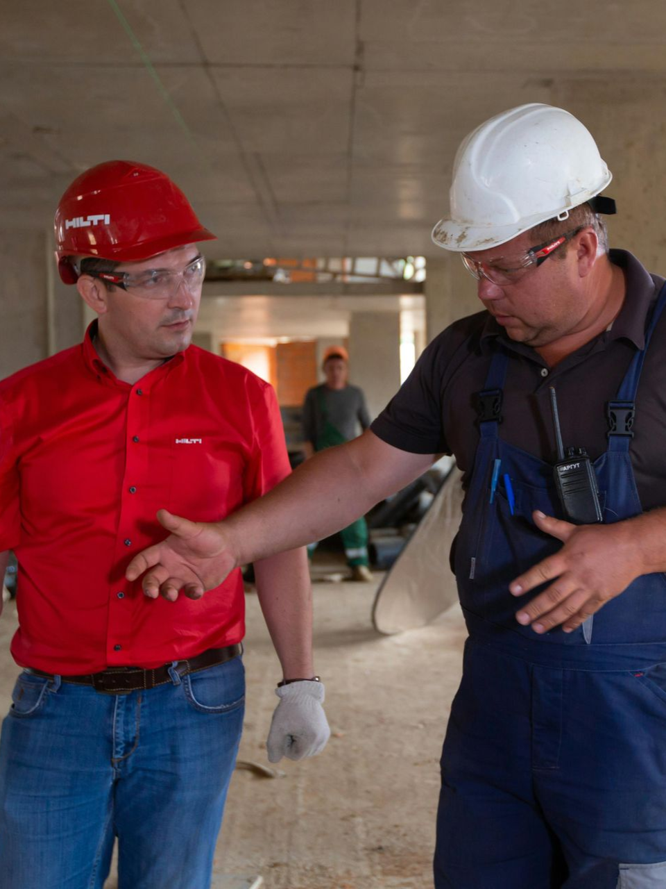 Two construction workers in hard hats talk inside a building under construction.