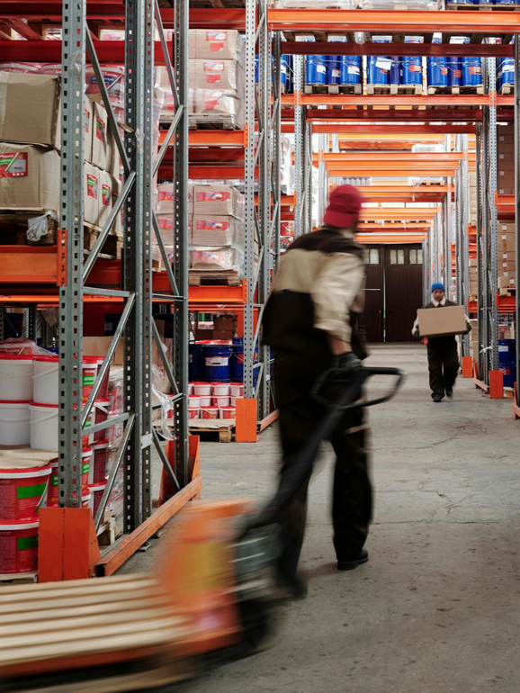 Workers moving pallets in a warehouse aisle with tall orange shelving and stacked goods.