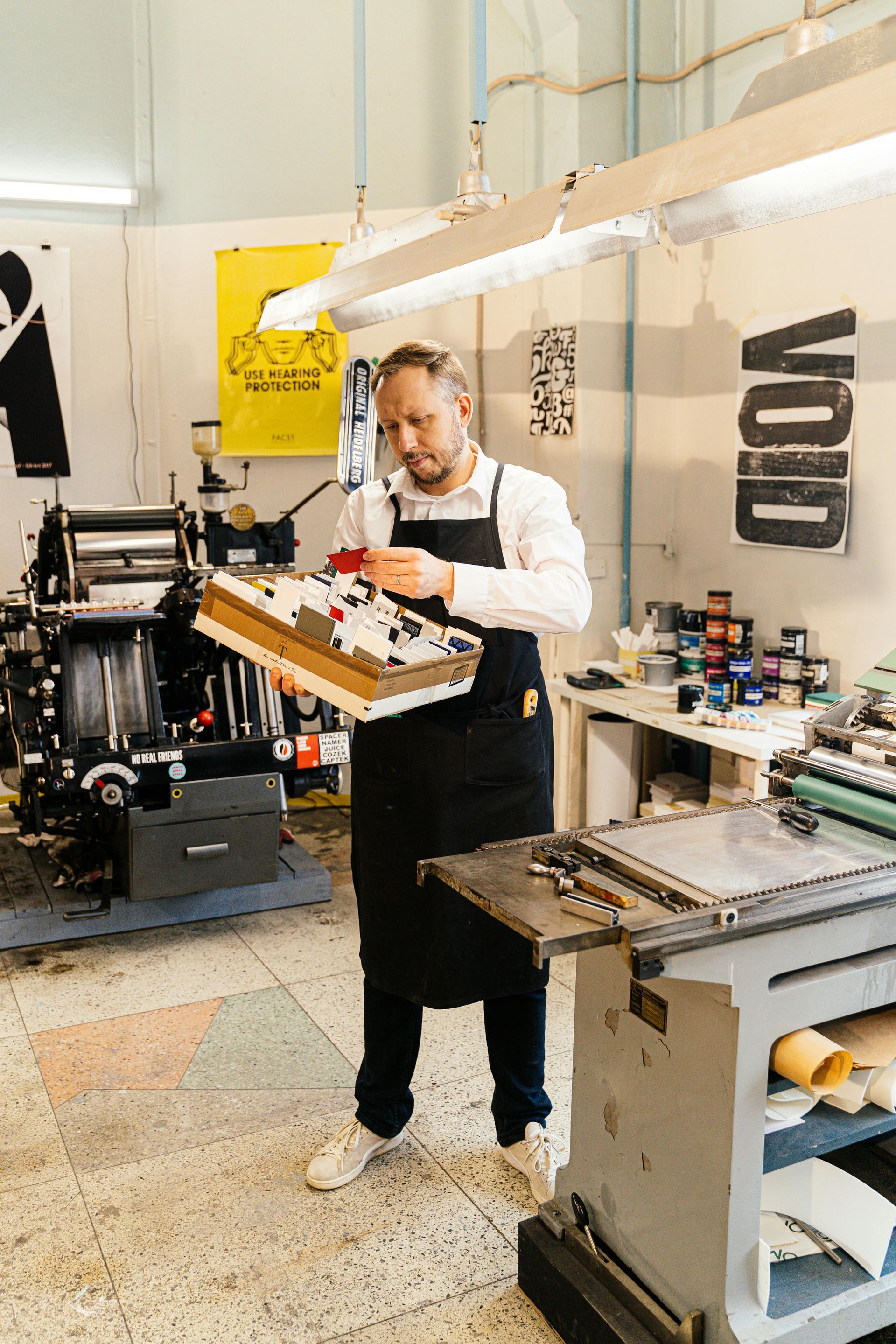 Person working in a print shop, holding a screen-printing frame beside a press.