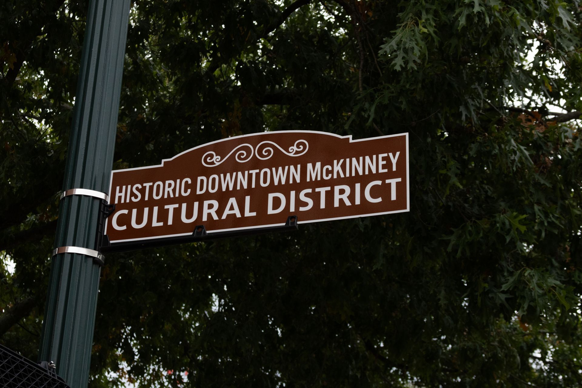 Sign for Historic Downtown McKinney Cultural District. Brown sign on a pole, against a background of green trees.