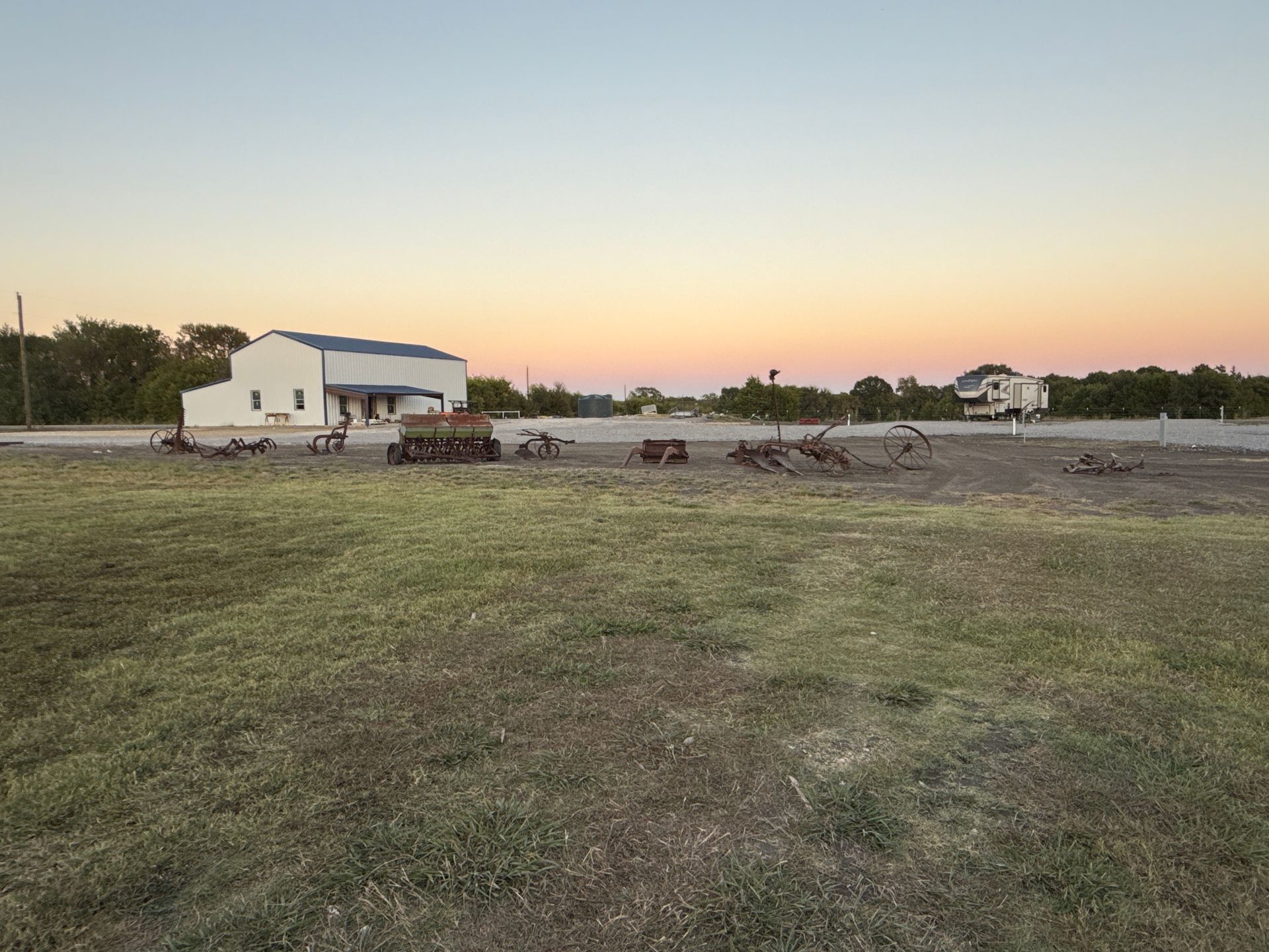 A building and vehicles sit on a grassy lot under an evening sky, with soft pink and orange hues.