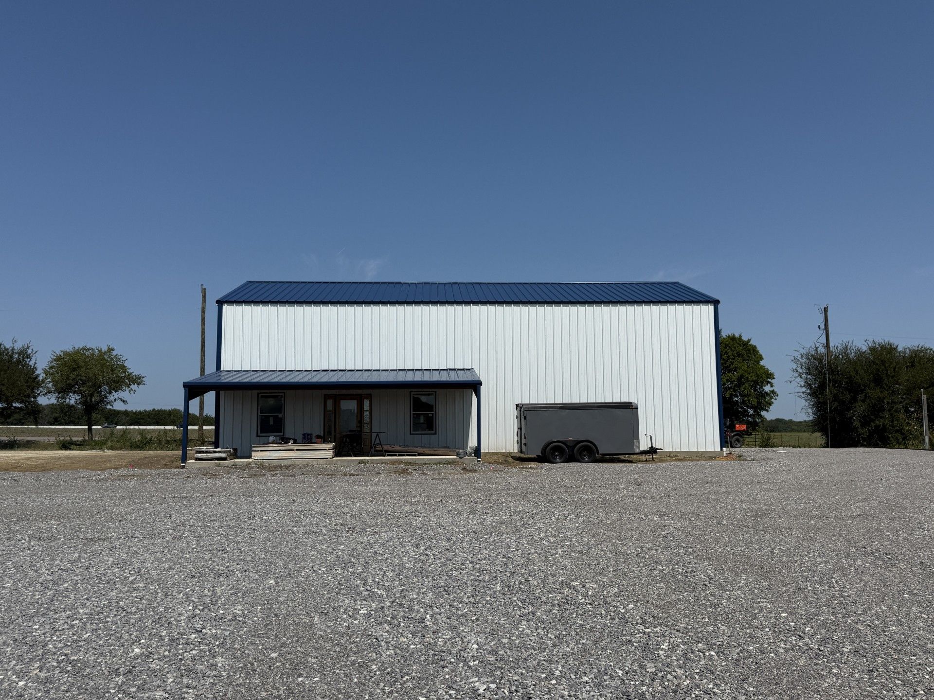 White metal building with blue roof and porch, parked trailer in front, gravel lot, clear sky.