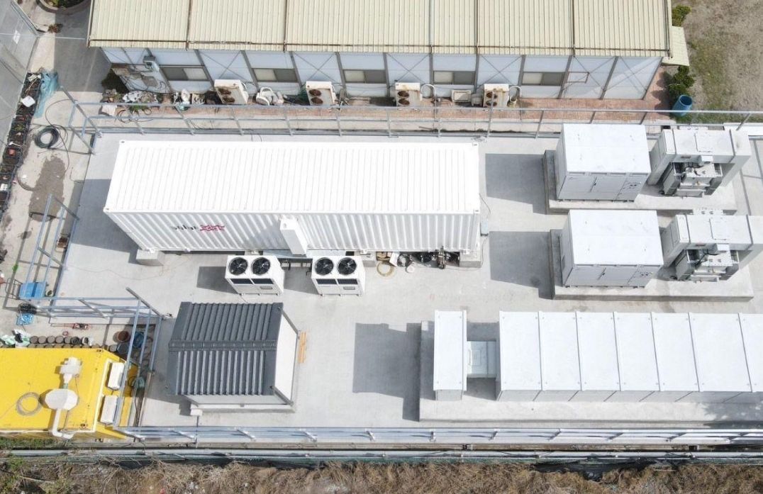 從上方俯瞰,可以看到一個白色電池儲能係統位於一棟裝有空調的建築物附近的混凝土平台上。