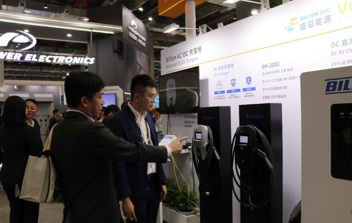 兩名男子在電動車充電站展台前指著設備。其他人則在展覽現場觀看。
