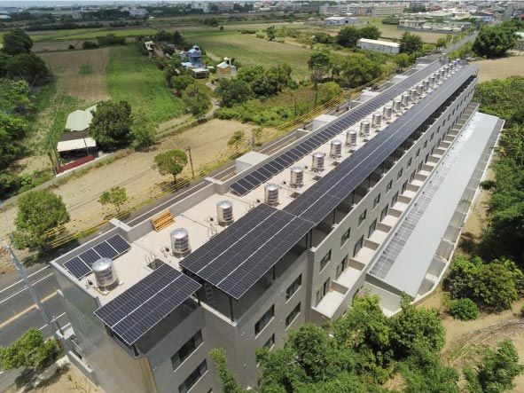 屋頂上安裝著成排的太陽能電池板，周圍環繞著綠樹和農田。