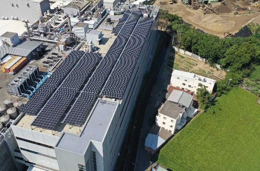 從空中俯瞰，一棟大型建築物的屋頂覆蓋著太陽能電池板，周圍環繞著其他建築物和綠植。