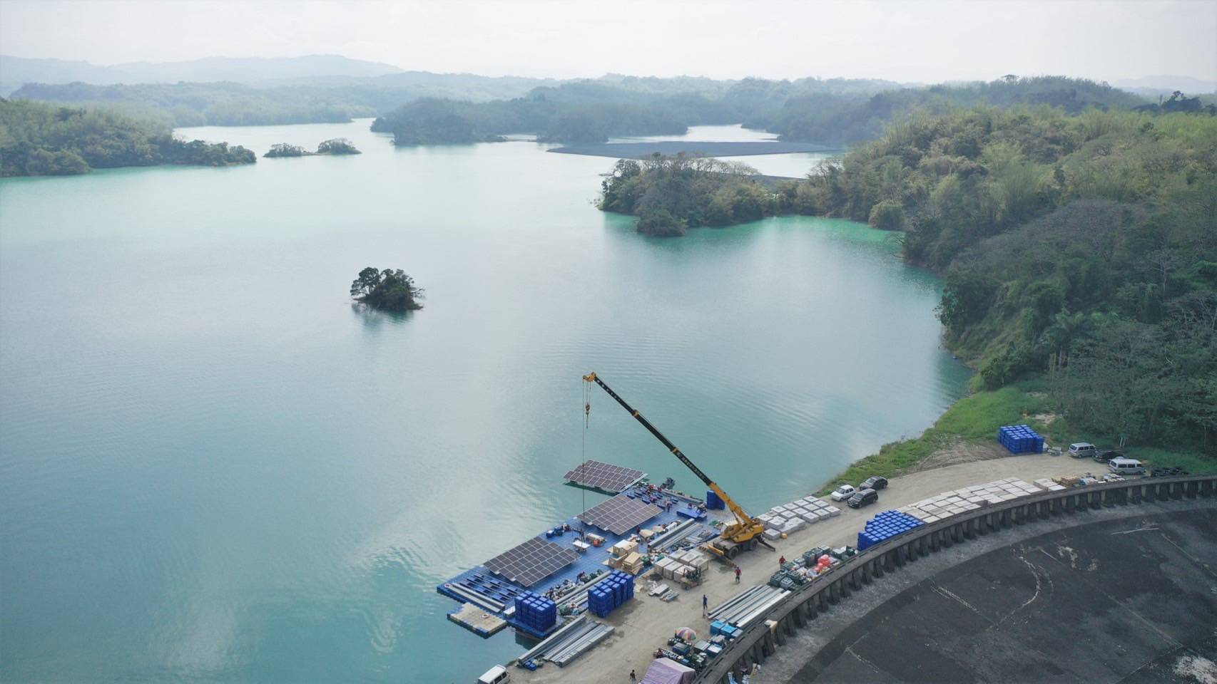 從空中俯瞰碧綠的湖泊，湖中點綴著小島，岸邊有一個建築工地，有起重機和設備。