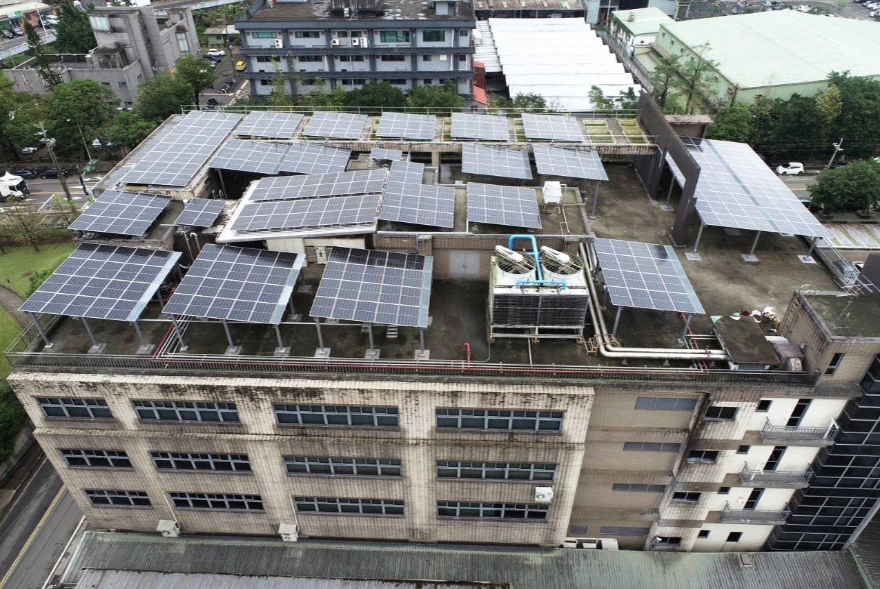 從高處俯瞰一座多層建築的屋頂，屋頂上覆蓋著太陽能電池板。背景中可以看到綠植和其他建築物。