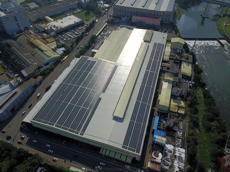 從空中俯瞰一座大型工業建築，屋頂上裝有太陽能電池板。該建築毗鄰街道、河流和其他建築物。