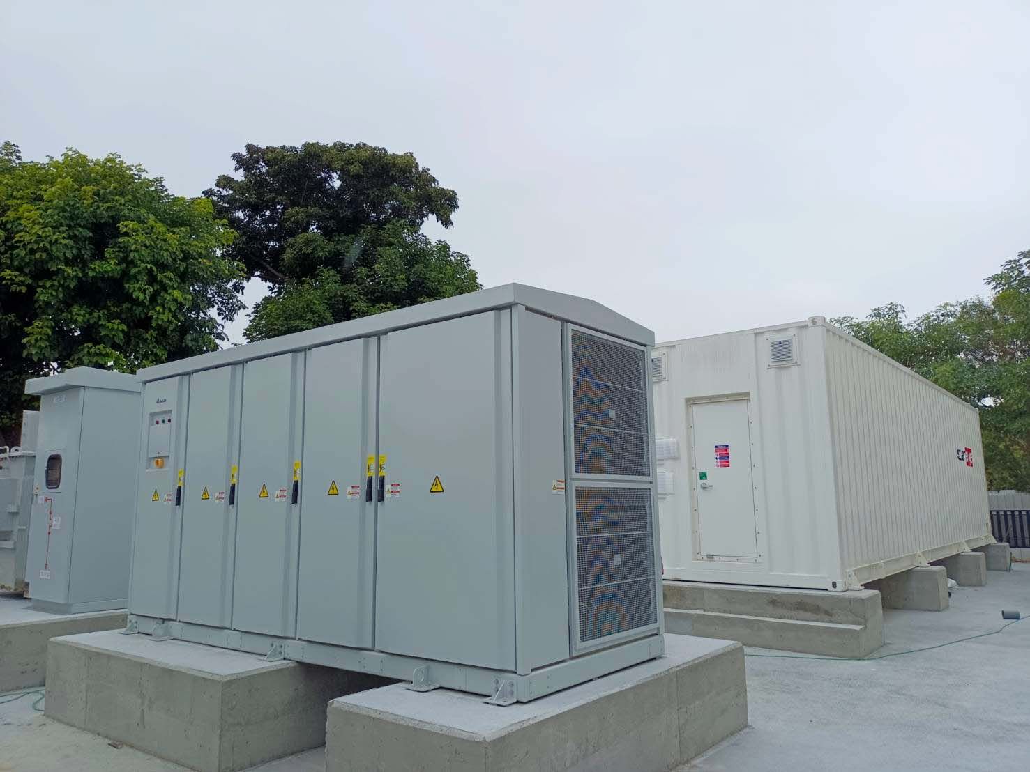 戶外電氣設備、灰色櫃子和白色容器，放置在混凝土塊上，背景是樹木。