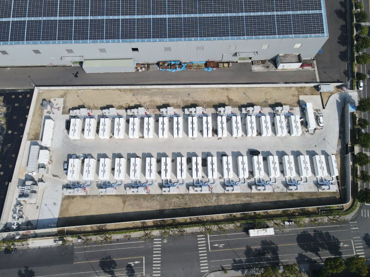 從空中俯瞰一座電池儲能設施，可以看到許多白色貨櫃。背景中可以看到一棟裝有太陽能板的建築物。
