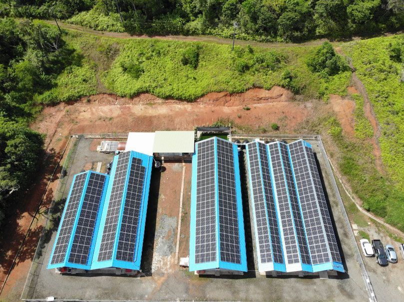從高處俯瞰一座太陽能發電站，藍色屋頂的建築物上裝有太陽能板，周圍環繞著綠色植被。