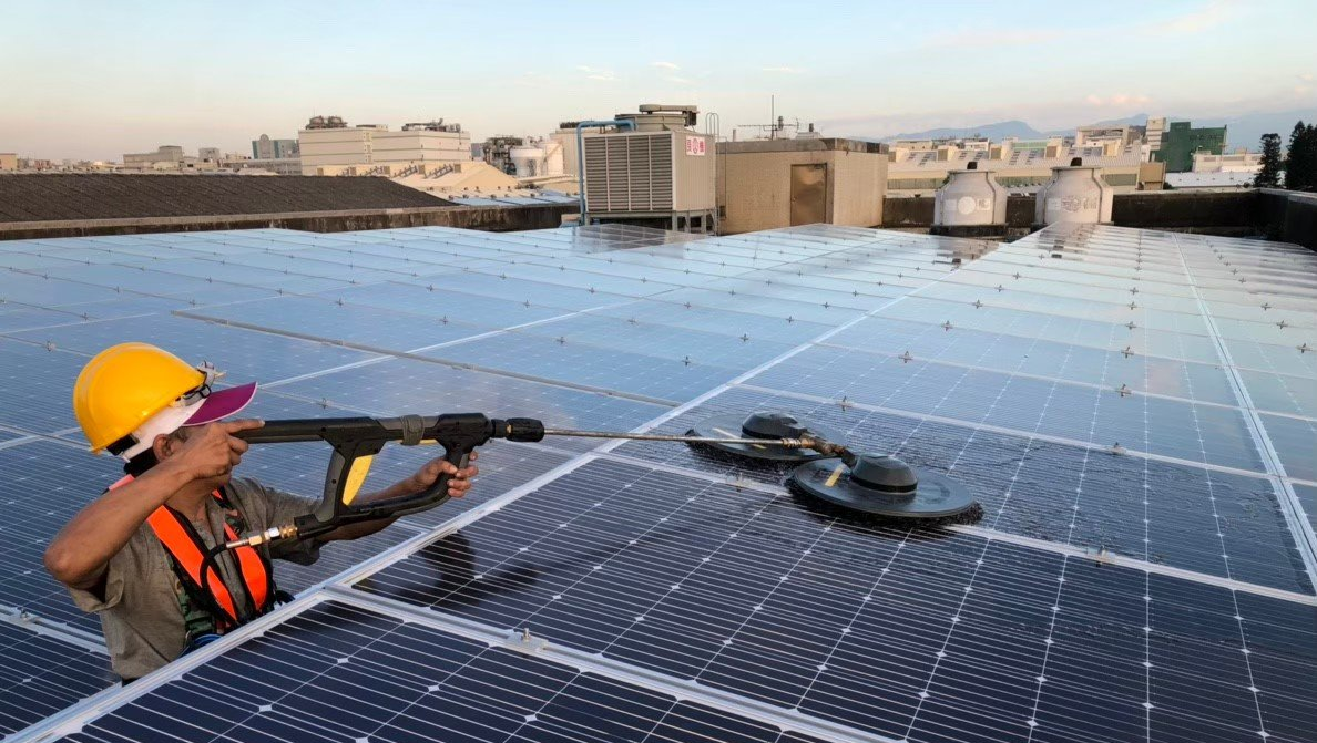 一名身穿安全服的人員正在屋頂上使用電動工具清潔太陽能板。背景是城市天際線。