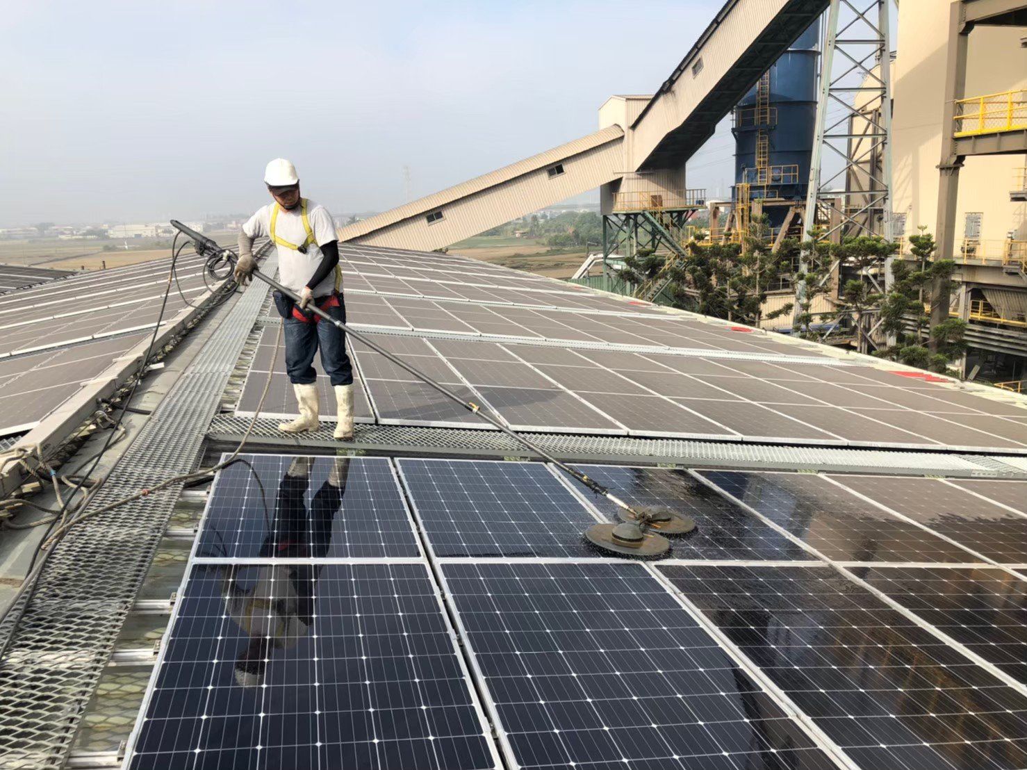 一名工人在屋頂上清潔太陽能電池板，背景中可以看到工業設備。