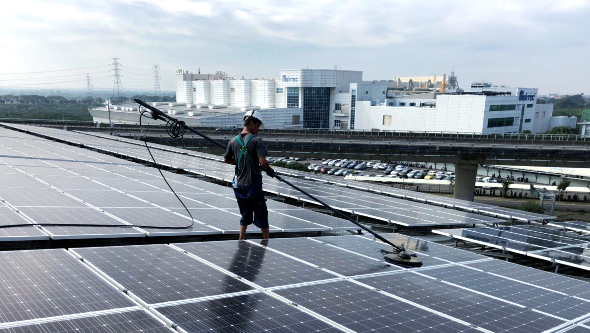 一名男子正在用長柄刷子清潔屋頂上的太陽能電池板，背景是一棟大型白色建築和陰沉的天空。