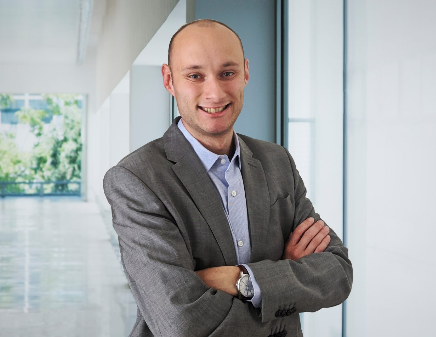 Un homme en costume gris, les bras croisés, souriant, dans un bureau lumineux.