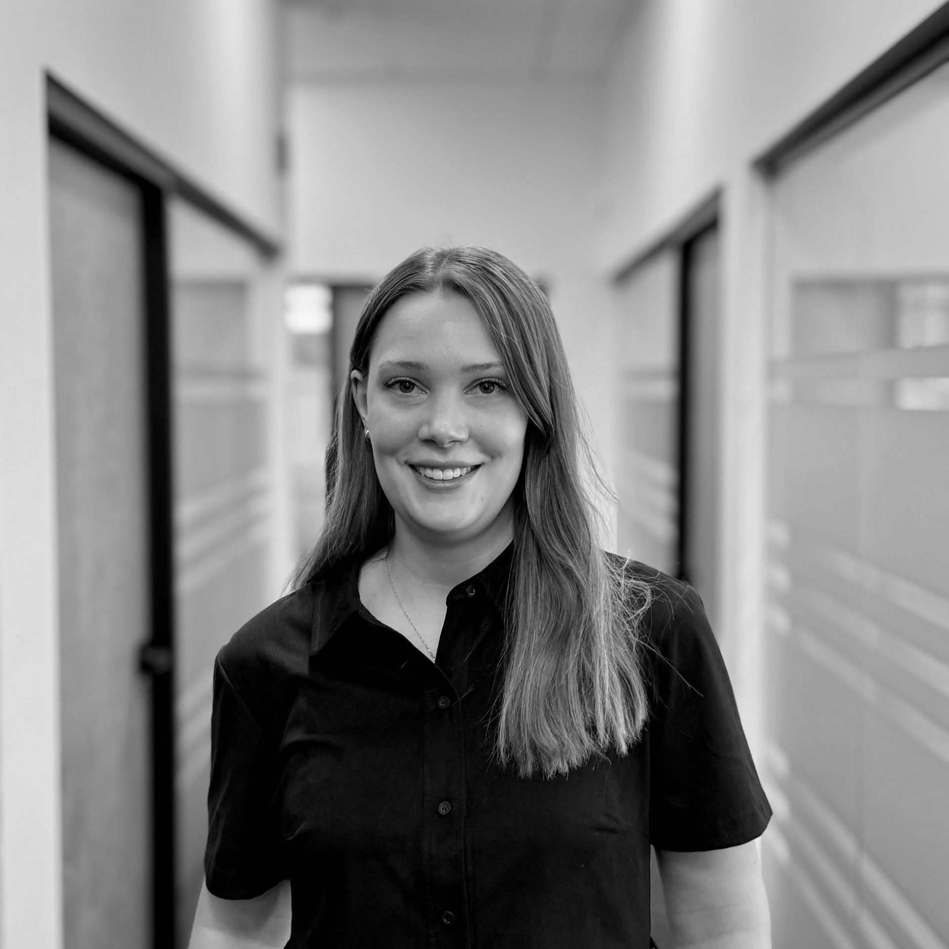 Femme en chemise noire souriante, debout dans un couloir de bureau.