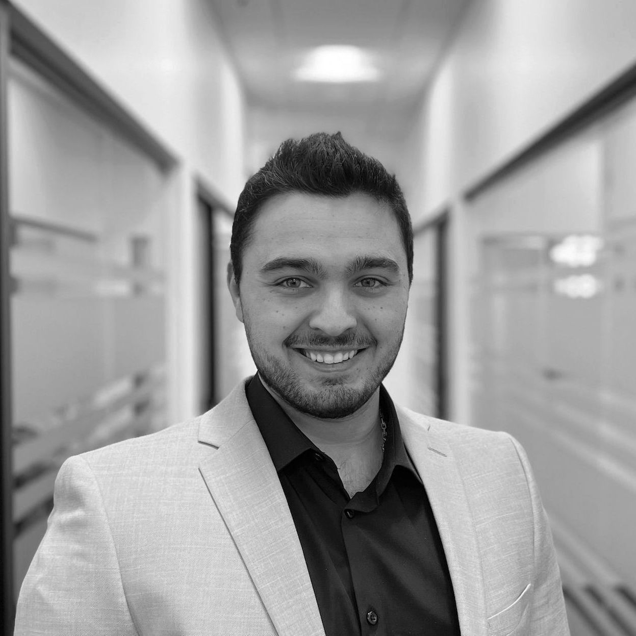 Un homme en blazer et chemise noire sourit dans un couloir de bureau.