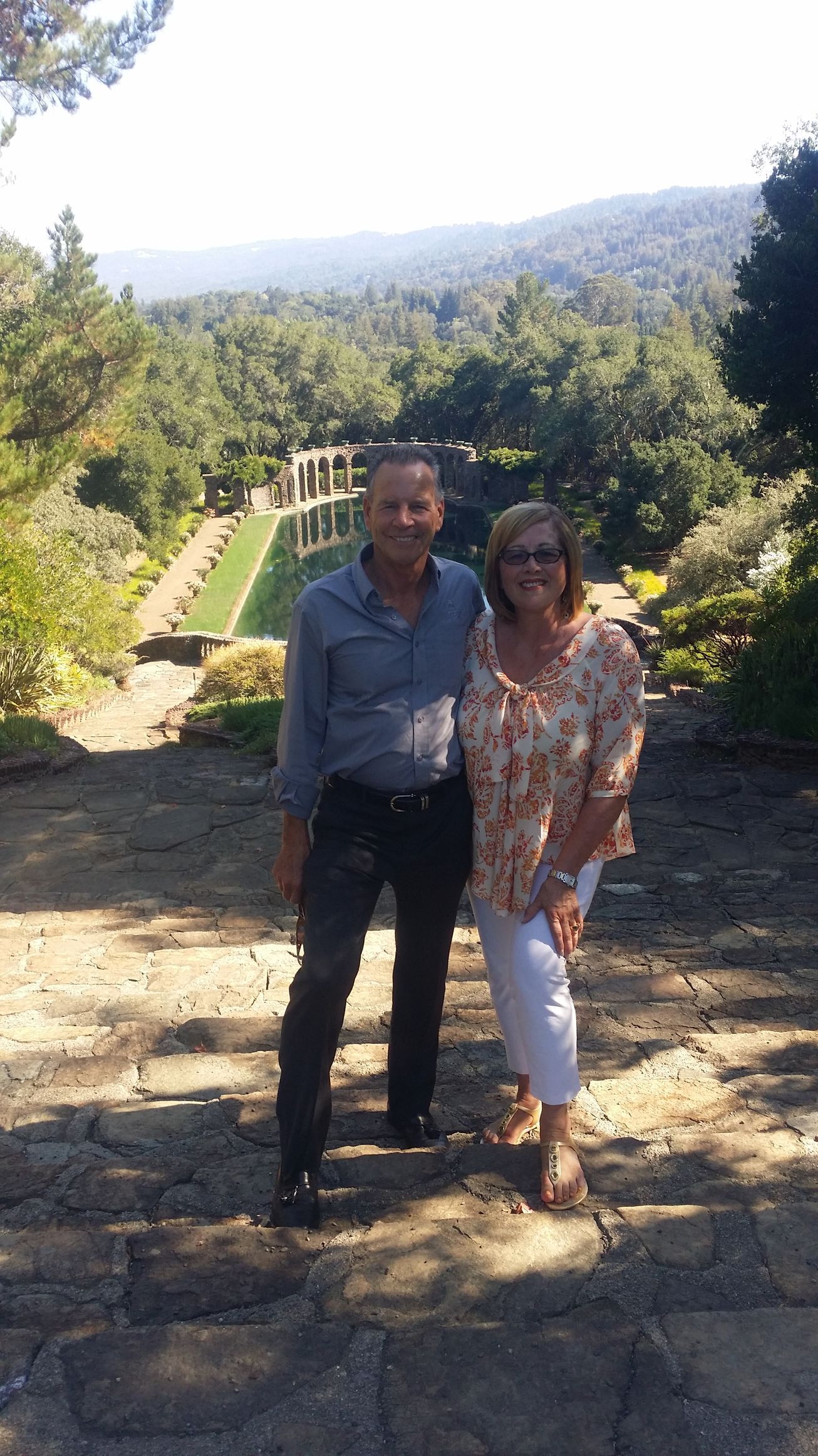 Two people standing on a stone path with lush green hills and a sunny scenic valley behind them