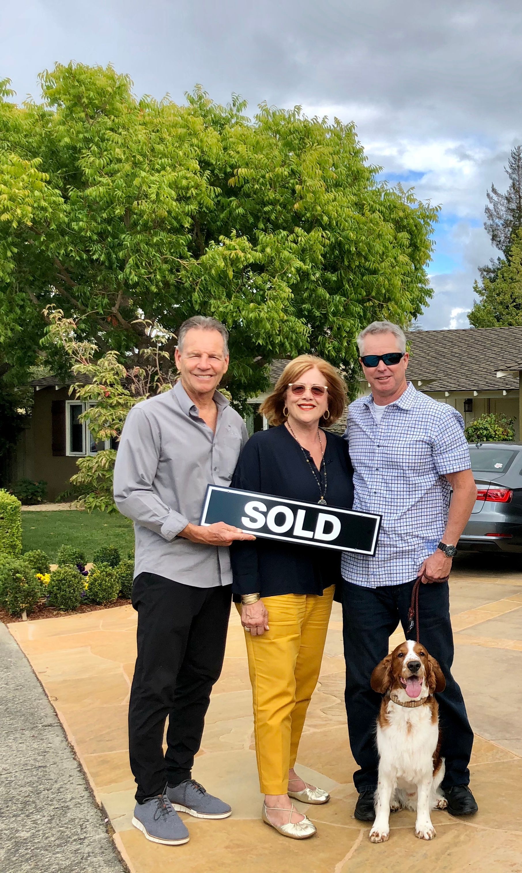 Three people hold a SOLD sign in front of a house, with a dog sitting beside them.