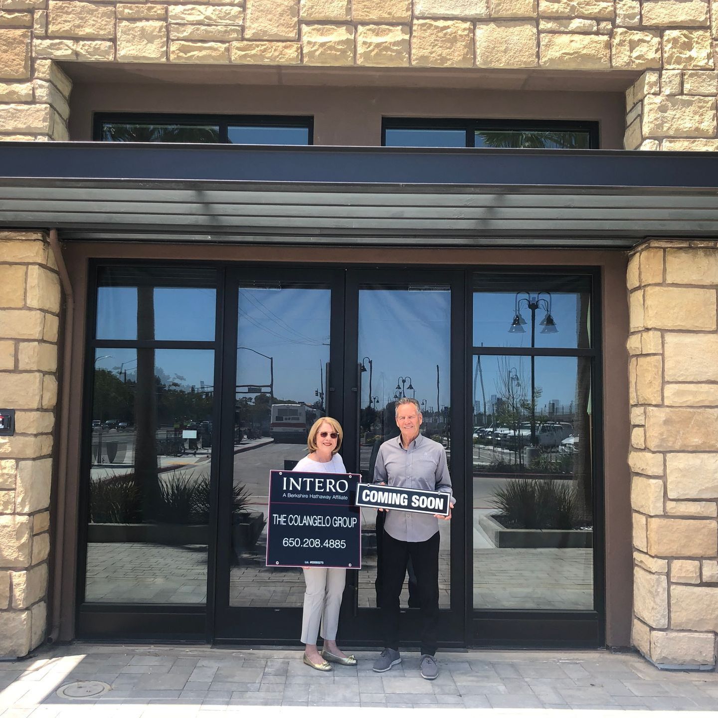Two people stand outside a glass storefront holding a real estate sign.