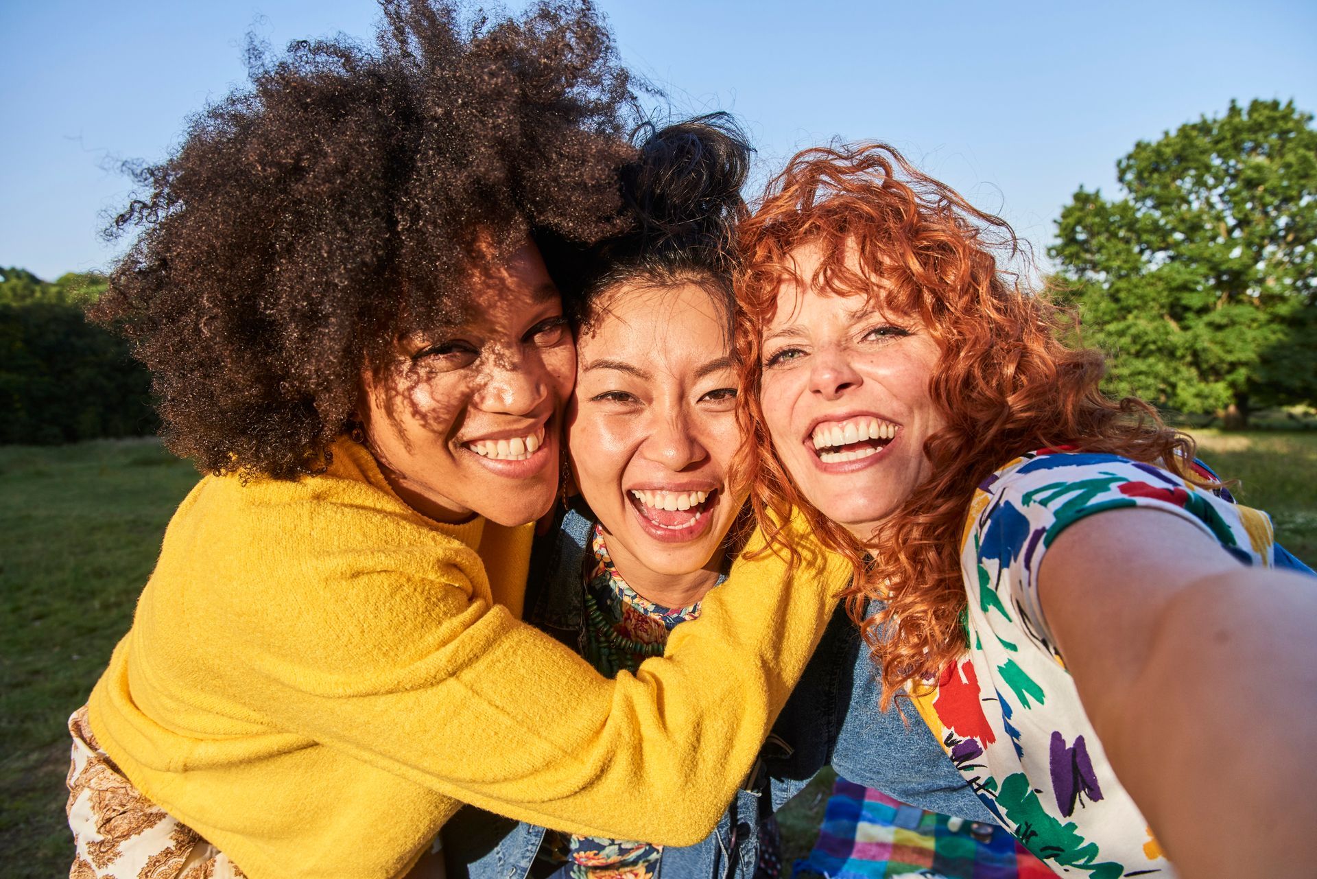 Three women are hugging each other and taking a selfie in a park.