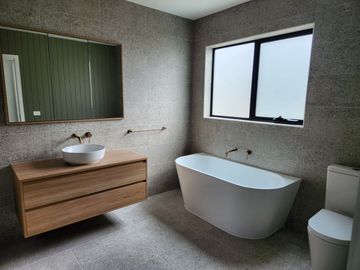 Bathroom with a white tub, toilet, wooden vanity, a mirror, and a window. Gray speckled walls.