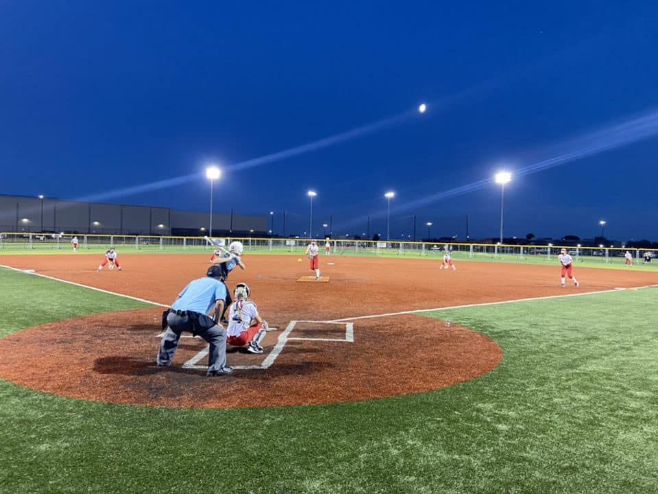 Youth baseball game at dusk, batter at home plate with catcher and umpire on a lit field.