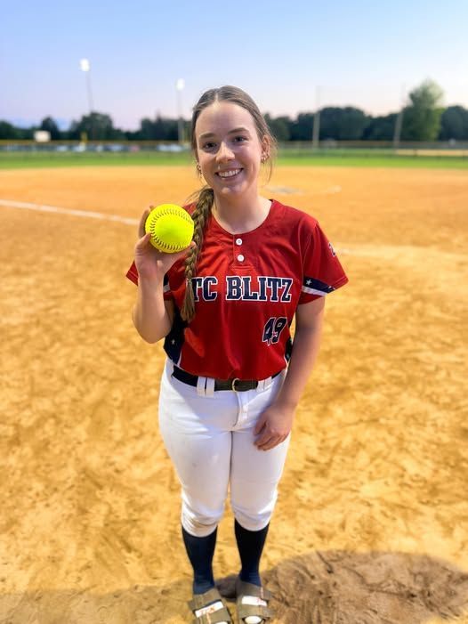 Youth softball player in red jersey and white pants holding a yellow ball on a dirt field.