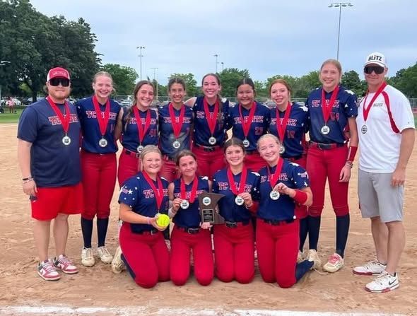 Softball team in red and blue uniforms posing with medals and a trophy on a field