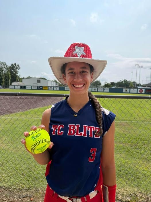 Smiling softball player in a red-and-white cowboy hat holds a neon yellow ball on the field.
