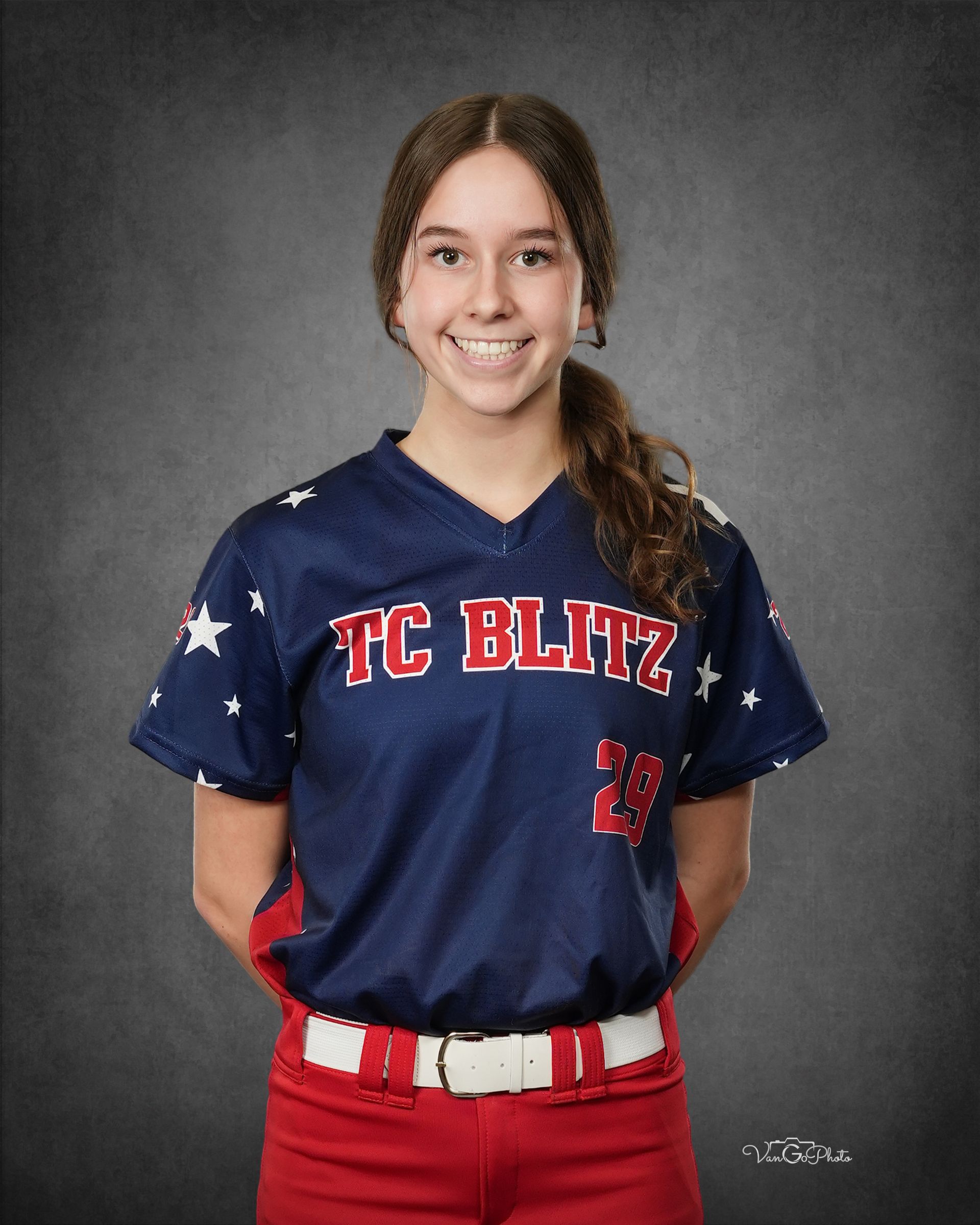Young softball player in navy and red uniform, smiling in a studio portrait against a gray background.
