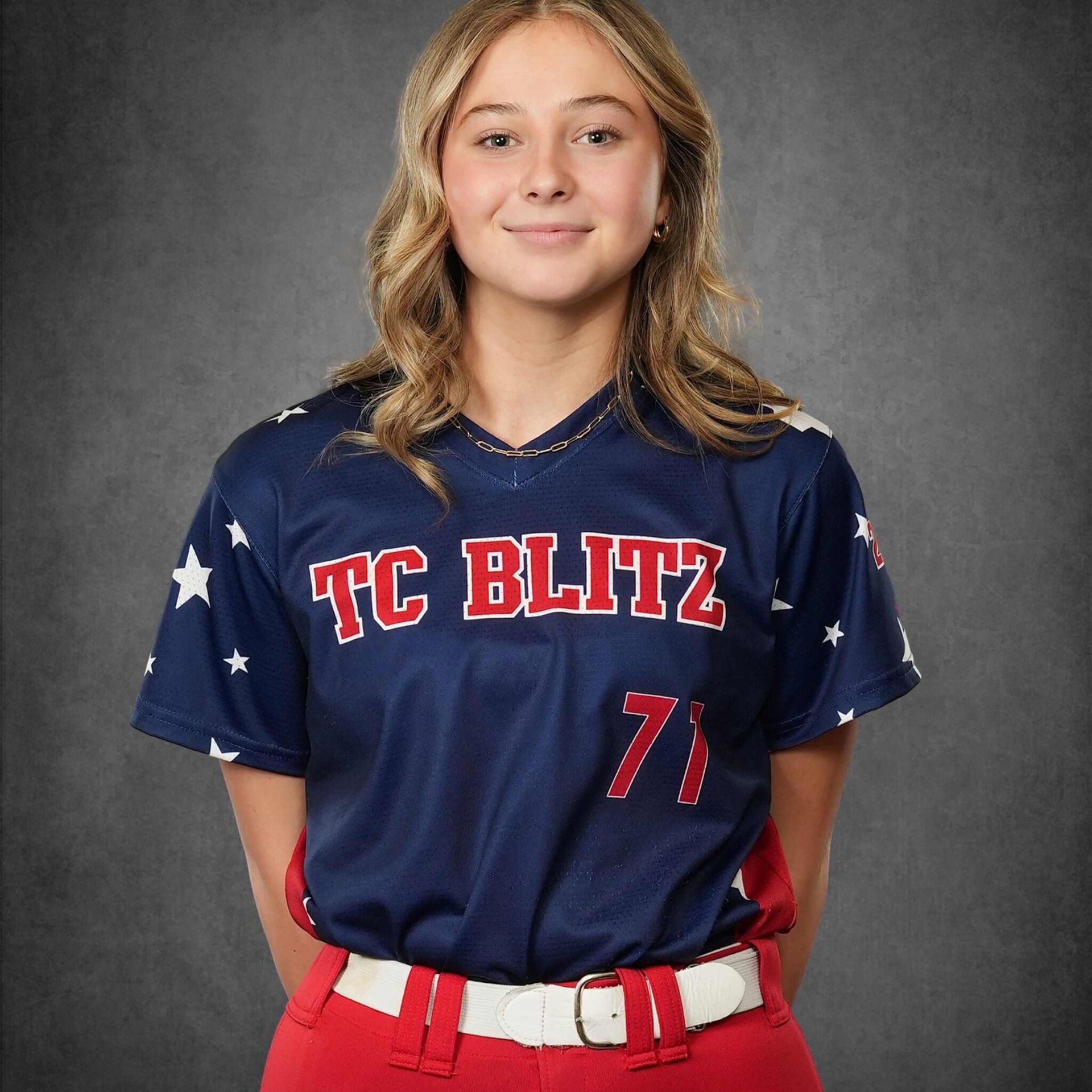 Youth softball player in navy TC BLITZ jersey with red pants, posing against a gray studio backdrop