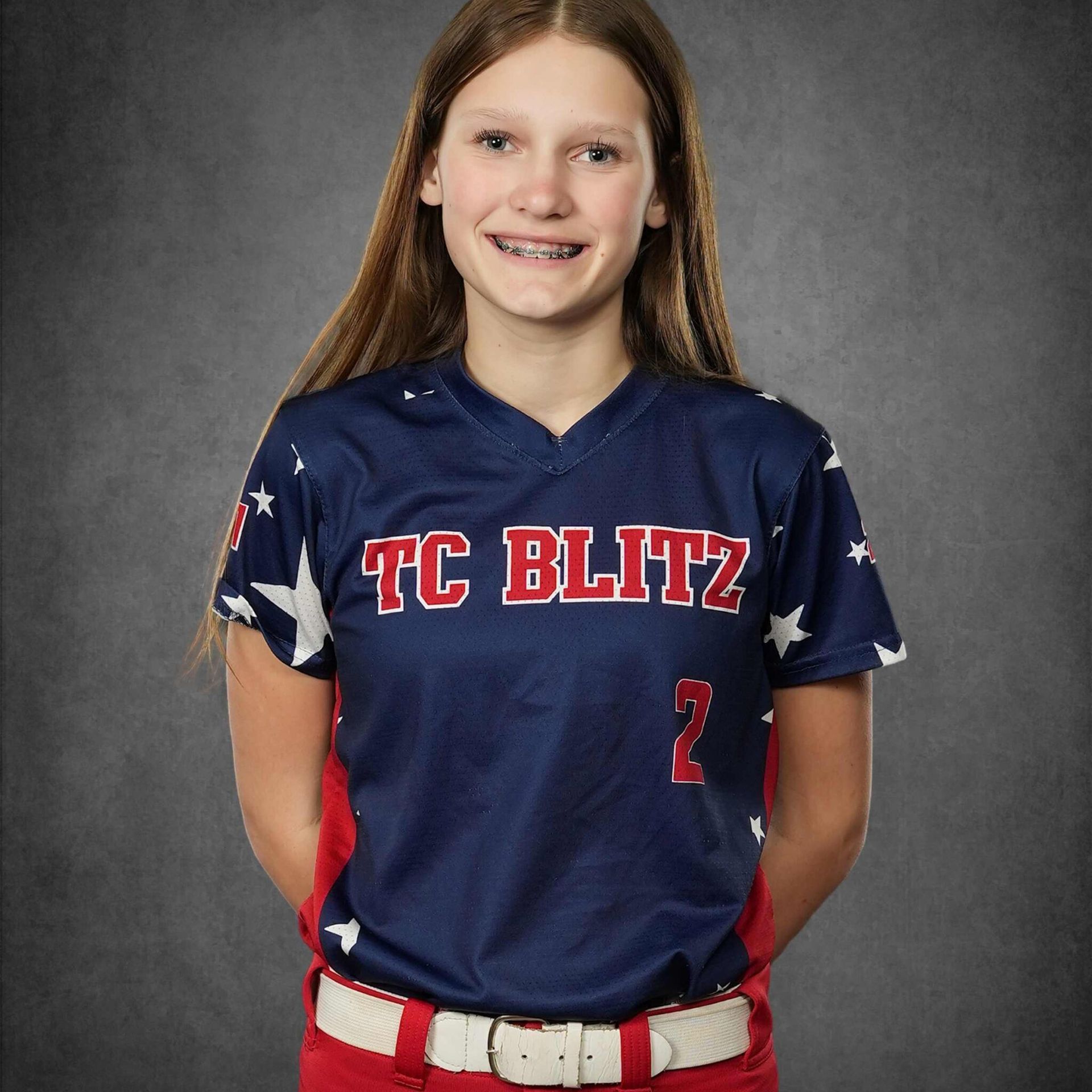 Young softball player in navy TC BLITZ jersey and red belt, smiling against a gray studio backdrop
